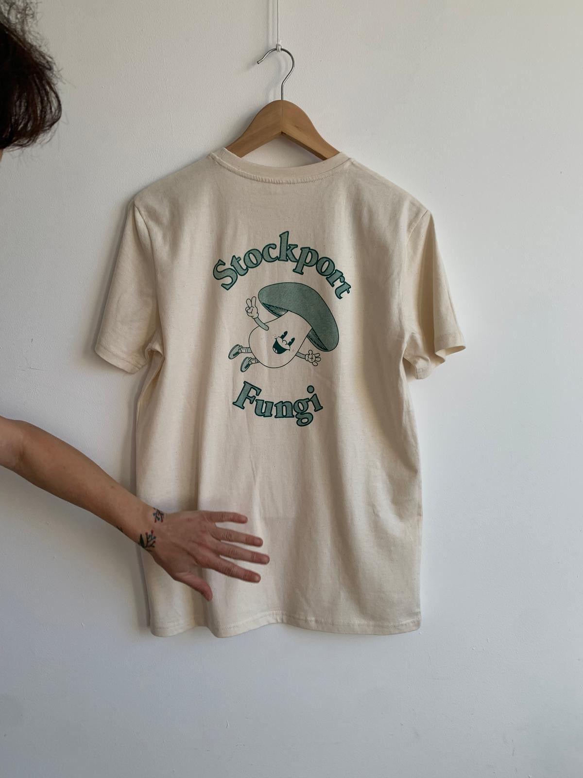 A beige t-shirt with 'STOCKPORT FUNGI' printed on the front, displayed on a wooden hanger against a white wall.